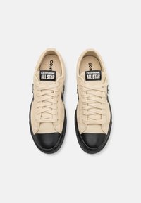 Beige kangaskengät mustalla kumisella varpaalla, mustilla yksityiskohdilla ja litteillä nauhoilla. Varustettu brändäyksellä kielellä ja sivussa.