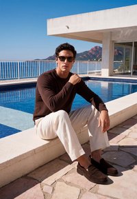 Homme portant des lunettes de soleil, un pull marron, un pantalon blanc et des mocassins foncés, assis au bord d'une piscine avec vue sur l'océan et les montagnes en arrière-plan.