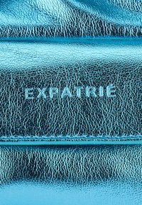 Metallic turquoise leather surface with a textured finish and creases. The brand name "EXPATRIE" is embossed in light blue at the center.