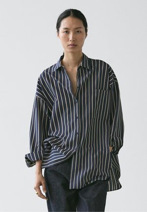 Kvinde med mørkt hår iført en oversized navyblå skjorte med hvide lodrette striber og mørke bukser, stående mod en ensfarvet lys baggrund.