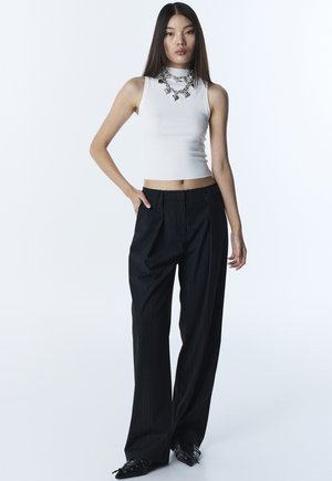 Giovane donna con canottiera bianca senza maniche, collana spessa d'argento, pantaloni neri a righe larghe e scarpe nere a punta, in piedi con una mano in tasca.