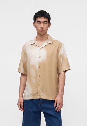 Jeune homme portant une chemise beige et blanche à manches courtes boutonnée et un jean bleu, debout devant un fond blanc uni.