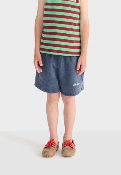Enfant portant un débardeur à rayures vertes et bordeaux, un short bleu avec le texte "Bobo Choses", et des chaussures beige à lacets rouges, debout devant un fond blanc.