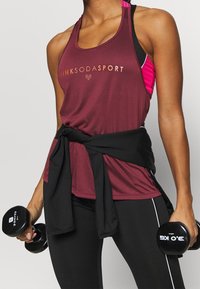 Mörkröd idrottstopp med guldbranding, kombinerad med svarta högmidjade leggings och svarta hantlar. Lätt textur, racerbaksdesign.