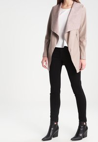 Chaqueta de cuero sintético beige con un amplio collar drapeado y cremallera lateral, combinada con pantalones negros de corte ajustado y botas de tobillo.