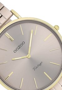 Montre en métal avec un boîtier doré, un cadran gris, des index fins et des aiguilles jaunes. Présente "OOZOO" et "Vintage" imprimés sur le cadran.