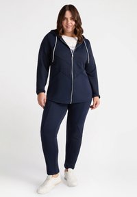 Felpa con zip navy e pantaloni abbinati, realizzati in tessuto testurizzato, con cappuccio regolabile e dettagli in pannelli a contrasto. Indossati con sneakers bianche.