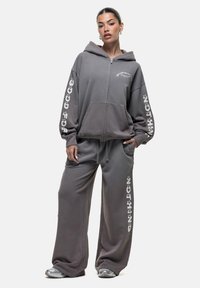 Grijze zip-up hoodie en bijpassende joggingsbroek met een oversized pasvorm, zachte textuur, zijzakken en witte logo-tekst op de mouwen en broek.