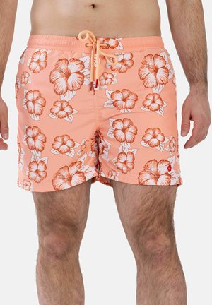 Mann trägt pfirsichfarbene Badeshorts mit orange-weißem Hibiskusblumenmuster und Kordelzug in der Taille, steht mit entspannt an den Seiten hängenden Armen.