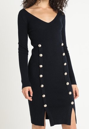 Femme portant une robe en maille côtelée noire à manches longues avec des boutons argentés disposés en deux rangées verticales avec des fentes au-dessus des genoux.
