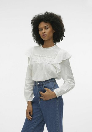 Vit långärmad blus med volanger vid axlarna och texturerad tyg, med spetsdetaljer. Parat med högmidjade blå denimjeans.