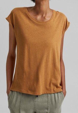 Woman wearing a short-sleeve, mustard-colored top and light gray pants, standing with hands in pockets against a plain gray background.