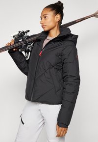 Doudoune noire avec capuche, présentant une surface texturée et des coutures diagonales. Soulignée par une fermeture éclair rouge et maintenue avec des skis.