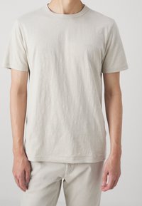 Helles beige T-Shirt mit kurzen Ärmeln, aus weicher Baumwolle, mit Rundhalsausschnitt und dezentem Logo auf der Brust, glatte Textur.