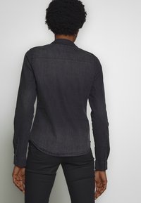 Dunkelblauses Denim-Hemd mit langen Ärmeln, abgerundetem Saum, Knopfmanschetten und subtiler Textur. Verfügt über eine Rückenpasse und minimalistische Details.