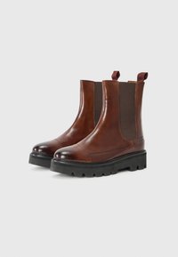 Bottines Chelsea en cuir marron avec des panneaux élastiques sur les côtés, bout rond et semelle épaisse à crans noire. Elles disposent de languettes pour un enfilage facile.