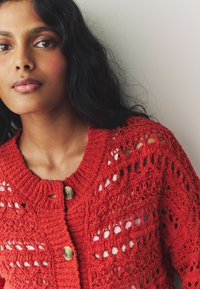 Cardigan au crochet rouge avec un design de mailles ouvertes, un col rond et deux boutons en bois. Modèle tricoté texturé avec des détails complexes.