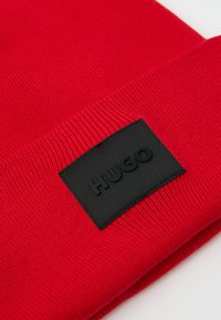 Rød strikket beanie med en foldet manchet, der har en rektangulær sort patch prægede med "HUGO". Glat tekstur, afslappet design.