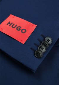 Marineblaues Sakko mit einem roten Etikett mit der Aufschrift "HUGO". Es hat vier schwarze Knöpfe an den Ärmeln und kontrastierende Nähte.