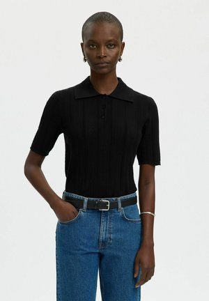 Femme aux cheveux courts portant un haut en tricot noir à manches courtes avec col, jean bleu, ceinture noire, boucles d'oreilles argentées en forme de créoles et bracelet, debout avec une main dans la poche.