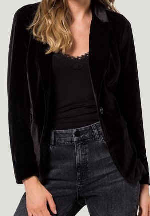 Woman wearing black velvet blazer, black lace-trimmed top, and high-waisted dark gray jeans against a plain background.