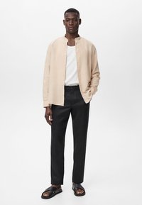 Beige linen shirt with a band collar, black trousers, and black sandals. Notable button details on the shirt. Simple, casual outfit.