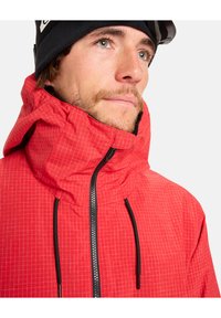 Rote Jacke mit Karomuster, hohem Kragen und Reißverschluss. Verfügt über schwarze Kordelzüge und einen strukturierten Stoff.