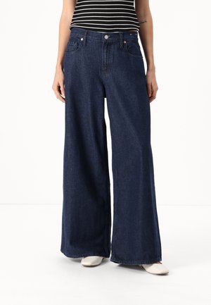 Osoba w ciemnoniebieskich szerokich dżinsach, białych butach i czarno-białej, bezrękawnikowej bluzce w paski, stojąca na jednolitym tle.