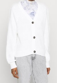 Cárdigan blanco de punto con cuello en V, que presenta cinco botones oscuros y puños acanalados. Acompañado de una blusa estampada y unos pantalones de mezclilla gris claro.