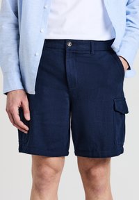 Bermuda cargo blu navy in tessuto testurizzato. Presenta tasche laterali e una chiusura con bottone in vita. Indossato con una camicia azzurra.