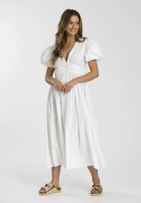 Weißes Midi-Kleid mit Puffärmeln, tiefem V-Ausschnitt, Knopfverschluss und geraffter Taille. Getragen mit beigen Sandalen.