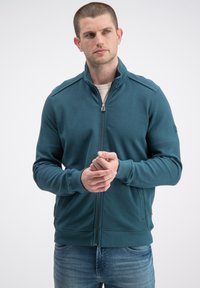 Turkusowa kurtka na zamek z teksturowanymi rękawami, ściągaczami przy mankietach i bocznymi kieszeniami. W połączeniu z jasną koszulą pod spodem. Sportowy fason.