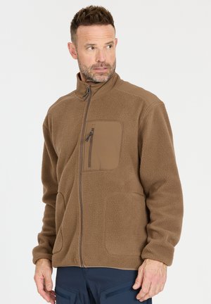 Mann trägt braune Fleecejacke mit Brust- und Handtaschen mit Reißverschluss, schaut nach links vor einem einfarbigen Hintergrund.