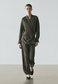 Frau in einem locker sitzenden dunkelbraunen Anzug mit Wickelblazer und weitem Hosenbein, trägt schwarze Ballerinas vor einfarbigem Hintergrund.