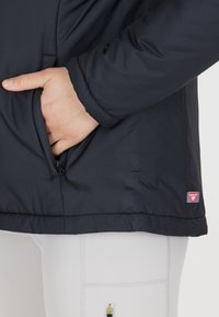 Doudoune noire avec une poche zippée visible et un logo Primaloft. Le tissu semble texturé et a une finition légèrement brillante.