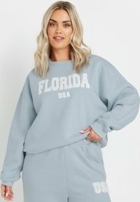 Sudadera de gran tamaño en azul claro con "FLORIDA USA" impreso en blanco, con puños acanalados y cuello redondo. Textura de tela suave visible.