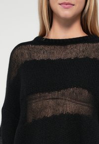 Schwarzer Strickpullover mit lockerem Gewebe und transparenten horizontalen Streifen. Dezente Textur mit weitem Ausschnitt und lässiger Passform.