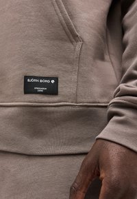 Κοντινό πλάνο σε τσέπη καφέ φούτερ με μαύρη ετικέτα Björn Borg που αναγράφει "Stockholm 1956" και ένα χέρι με σκούρο δέρμα ακουμπάει δίπλα της.