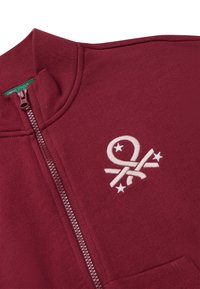 Sudadera con cremallera color burdeos y cuello alto, con bordado blanco de un logo y estrellas, y bolsillo delantero. Tela de textura suave.