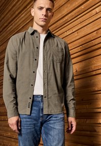 Olive green button-up shirt with a chest pocket, worn over a white t-shirt, paired with light blue jeans, set against a wooden background.