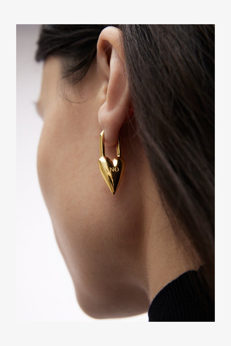 Auksiniai, širdelės formos auskarai su užrakto dizainu, kuriuose išraižytas žodis "NE". Lygus metalinis paviršius.