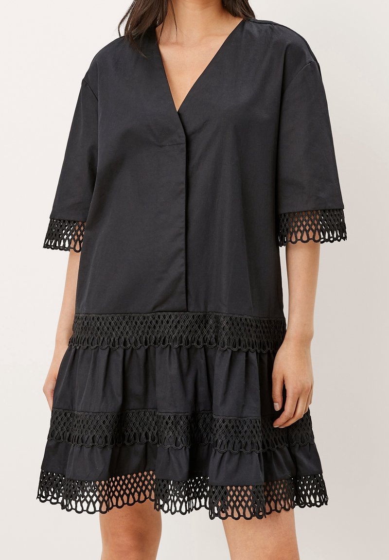 Femme portant une robe noire ample avec des manches courtes et une bordure en dentelle noire décorative aux manches, à la taille et à l'ourlet.