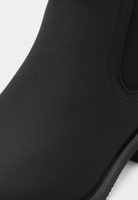Gros plan sur une botte noire en matériau mat lisse, couture visible sur le côté et panneau élastique noir.