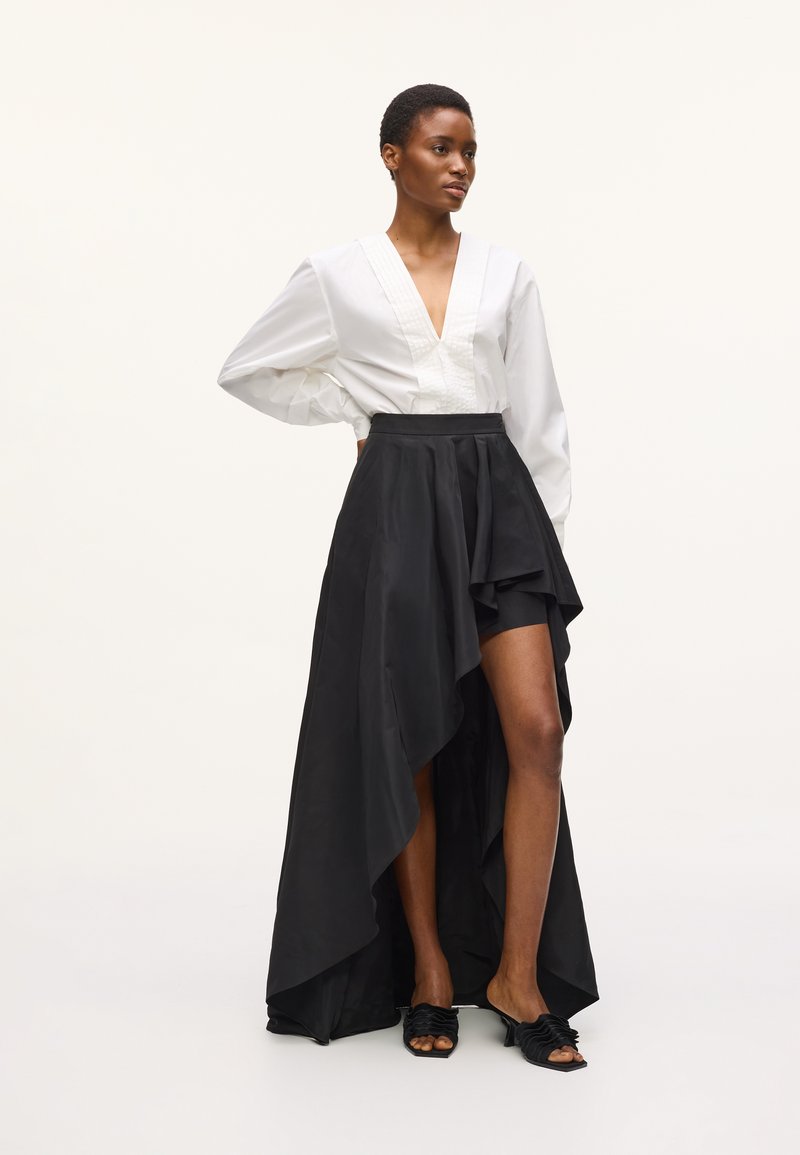 Γυναίκα με κοντά μαλλιά που φοράει λευκό πουκάμισο με βαθύ V και ασύμμετρη μαύρη φούστα με ψηλή σκίσιμο και μαύρα σανδάλια με τακούνι.