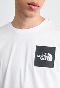 Weißes Baumwoll-T-Shirt mit einem schwarzen quadratischen Logo und dem Text "THE NORTH FACE" sowie einem Halbkreis-Design; kurze Ärmel, Rundhalsausschnitt.