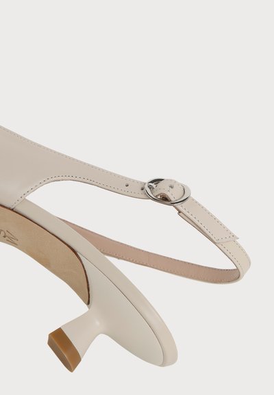 Gros plan sur une chaussure beige à petit talon, bout pointu, bride à la cheville et boucle argentée sur fond blanc uni.