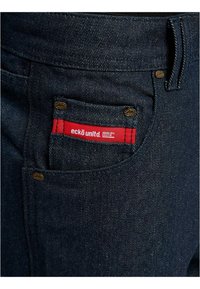 Tummansinisen farkkujen tasku punaisella etikettillä, jossa lukee "eckō unltd. DENIM DIV. RAW72" ja pienillä messinkinauloilla.