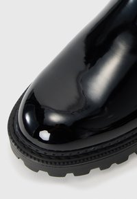 Botín de cuero charol negro con un acabado brillante, suela de goma gruesa y patrón de tracción robusto; punta redondeada y detalles de costura reforzada.