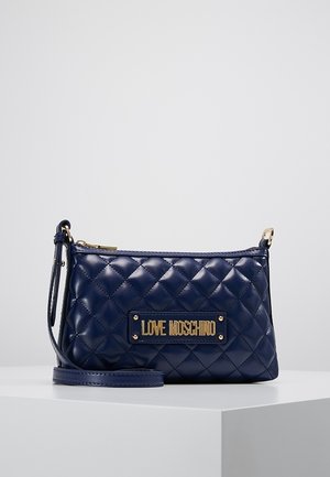 Granatowa pikowana skórzana torebka na ramię z złotą plakietką z logo "LOVE MOSCHINO" i zamkiem błyskawicznym, spoczywająca na białym piedestale.