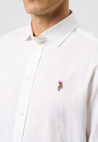 Valkoinen pellavapaita, jossa on napillinen kaulus ja pieni kirjailtu kuvio vasemmassa rinnassa. Kangas on teksturoitua kudontaa.